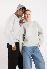 Két modell világosszürke, oversized pulóverben, bordás mandzsettával, fekete és kék farmert viselve. Az egyiküknél szürke sapka van.