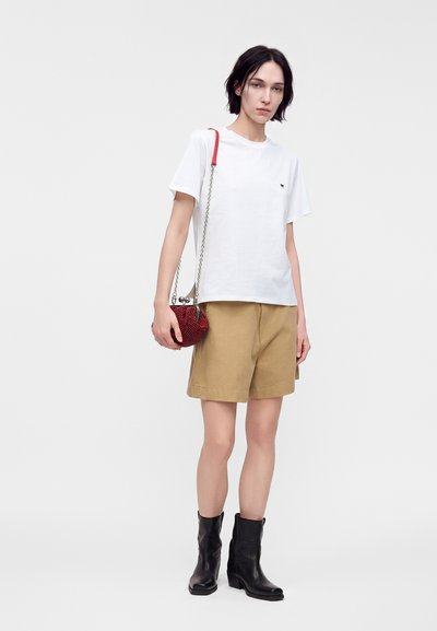 Femme portant une chemise blanche à manches courtes, un short beige à longueur genou, des bottines noires, tenant un petit sac à main rouge avec une bandoulière chaîne.