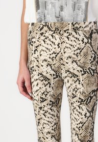 Beige leggingi z vzorcem kače z gladko teksturo, ki vsebujejo črno-bele vzorce. Prilegajoča zasnova z srednje visokim pasom.