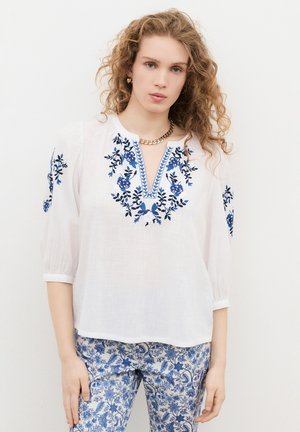 Donna con capelli ricci indossa una camicetta bianca con ricamo floreale blu e pantaloni con motivo floreale blu, in piedi davanti a uno sfondo bianco semplice.