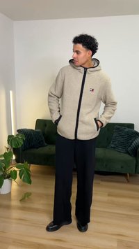 Beežika villasegu jope kapuutsiga, tõmblukuga ja tumelilla äärisega. Mustad laiade säärtega püksid ja mustad kingad. Siseinterjöör rohelise diivaniga.