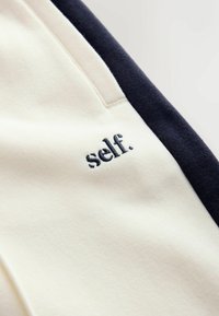 Kreminės ir tamsiai mėlynos spalvos audinys su tekstūruotu paviršiumi, ant kreminės dalies tamsiai mėlynomis siūlėmis išsiuvinėtas žodis „self.“. Matomos kišenės.