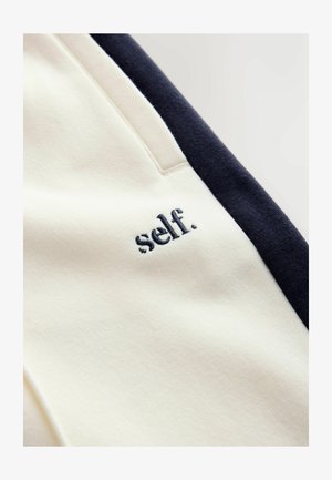 Krémová a námornícka tkanina s textúrou, na krémovej časti vyšitý nápis "self." v námorníckej farbe. Viditeľné vrecká.