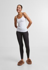 Débardeur blanc ajusté, leggings noirs et pantoufles en peluche brunes. Le mannequin se tient avec les bras derrière le dos, souriant, sur un fond uni.
