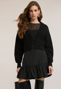 Musta, lyhyt neuletakki teksturoidulla neuloksella, jossa on syvä V-pääntie ja pitkät hihat. Päällä musta liehukehelmainen mekko.