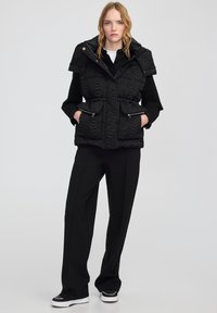 Czarna pikowana kamizelka z kapturem, z teksturowaną powierzchnią, dwiema kieszeniami na zamek i dopasowanym krojem, w połączeniu z czarnymi spodniami i trampkami.