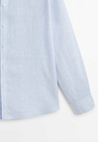 Chemise bleu clair à manches longues avec fines rayures blanches verticales et boutons blancs, manchette visible sur fond blanc uni.