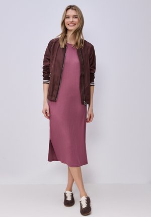 Hymyilevä nainen pukeutuneena purjeriviseen violettiin midimekkoon, jossa on sivuhalkaisija, ruskea bomber-takki ja ruskeat tennarit valkoisilla nauhoilla, seisoo yksivärisen taustan edessä.