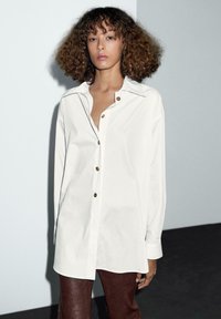 Camicetta bianca con colletto e chiusura frontale a bottoni, con maniche lunghe e un leggero lucido, indossata sopra pantaloni in pelle marrone.