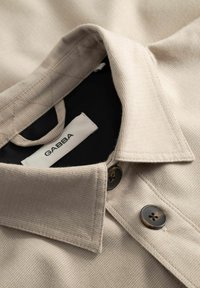 Col de chemise en tissu beige texturé avec deux boutons marron et une étiquette blanche portant la mention "GABBA" à l'intérieur de l'encolure.