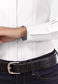 Chemise blanche à col classique avec des poignets boutonnés, associée à une ceinture en cuir noire avec une boucle en argent, portée sur un jean en denim bleu foncé.