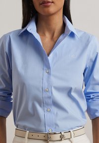 Ljusblå skjorta med knappar framtill, klassisk krage, bröstficka och uppvikta ärmar. Har vita knappar och diskret branding på bröstet.