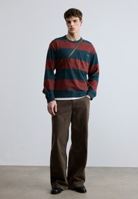 Μπλούζα με μακριά μανίκια και ρίγες σε βαθύ τιρκουάζ και μπορντό, με μια τσέπη στο στήθος. Συνδυασμένη με καφέ παντελόνι φαρδιάς γραμμής και μαύρα παπούτσια.