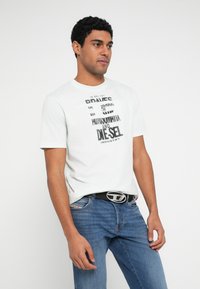 Camiseta gris claro con texto gráfico negro, combinada con vaqueros azules y un cinturón negro con hebilla plateada, estilizada de manera casual.