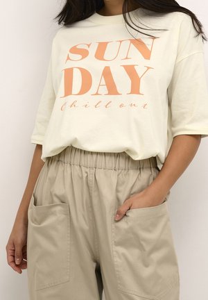 Młoda osoba nosząca kremową oversize koszulkę z napisem "SUNDAY chill out" oraz beżowe spodnie z wysokim stanem i dużymi przednimi kieszeniami.