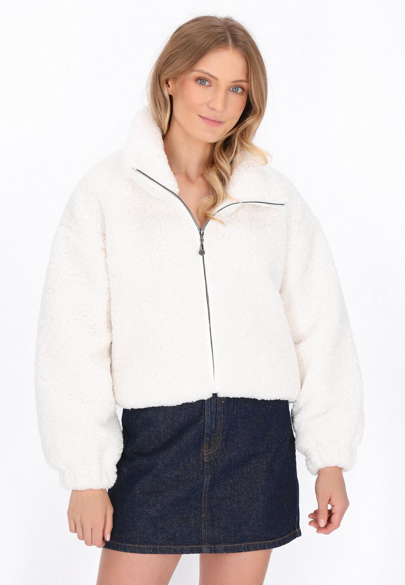 Bílá, chlupatá zipová bunda s vysokým límcem a oversized rukávy, kombinovaná s tmavými džíny sukní. Měkká textura a volný střih.