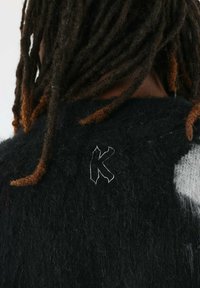 Pull en tricot noir avec des accents en fibres blanches, présentant une finition texturée. Un 'K' brodé en relief orne le col à l'arrière.