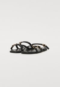 Sandales plates noires avec des straps fins, accents dorés et une semelle lisse. Le design est minimaliste avec des éléments décoratifs près de la cheville.