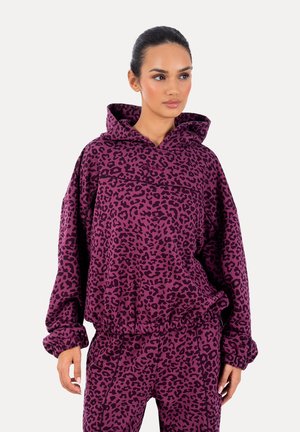 Kobieta ubrana w pasujący fioletowy komplet z kapturem i spodniami w panterkę, stojąca z rękami swobodnie opartymi po bokach, patrząca lekko w prawo.