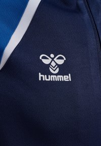 Gros plan sur un tissu bleu marine avec un détail de rayures blanches et bleues et un logo Hummel blanc présentant un design stylisé d'abeille et du texte.
