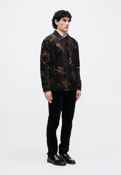 Jachetă din bumbac negru cu modele abstracte colorate, purtată împreună cu pantaloni negri și pantofi negri. Modelul stă drept pe un fundal simplu.