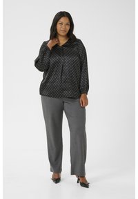 Blouse en satin noir à pois blancs, avec un devant boutonné et des manches longues, associée à un pantalon gris ajusté et des chaussures noires.