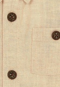 Camisa de lino beige con cuello clásico, bolsillo frontal y botones marrón oscuro con un diseño en relieve. Tejido texturizado con un entramado natural.