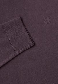 Gros plan sur une manche pliée et la zone de la poitrine d'un pull violet en maille avec un petit logo géométrique en relief.