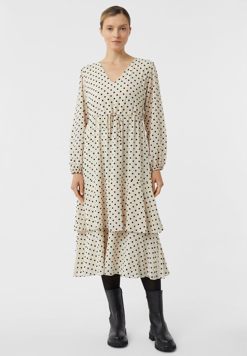 Sukienka z długim rękawem w kolorze kremowym w czarne groszki, z dekoltem w serek, marszczoną talią i falbanami na dole. Połączona z czarnymi botkami do kostki.