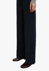 Pantaloni a gamba larga color blu navy realizzati in tessuto liscio, con polsino spesso e dettaglio sottile di piega. Abbinati a scarpe marrone scuro.