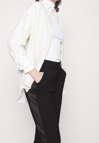 Licht crèmekleurig button-up shirt over een gestructureerde witte top, gecombineerd met zwarte broeken met een satijnen zijstreep en zijzakken.