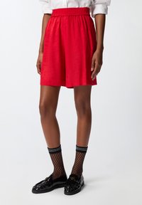 Shorts rossi con vita elastica, caratterizzati da una texture liscia e un motivo sottile. Abbinati a calze di rete nere e mocassini neri con un logo.