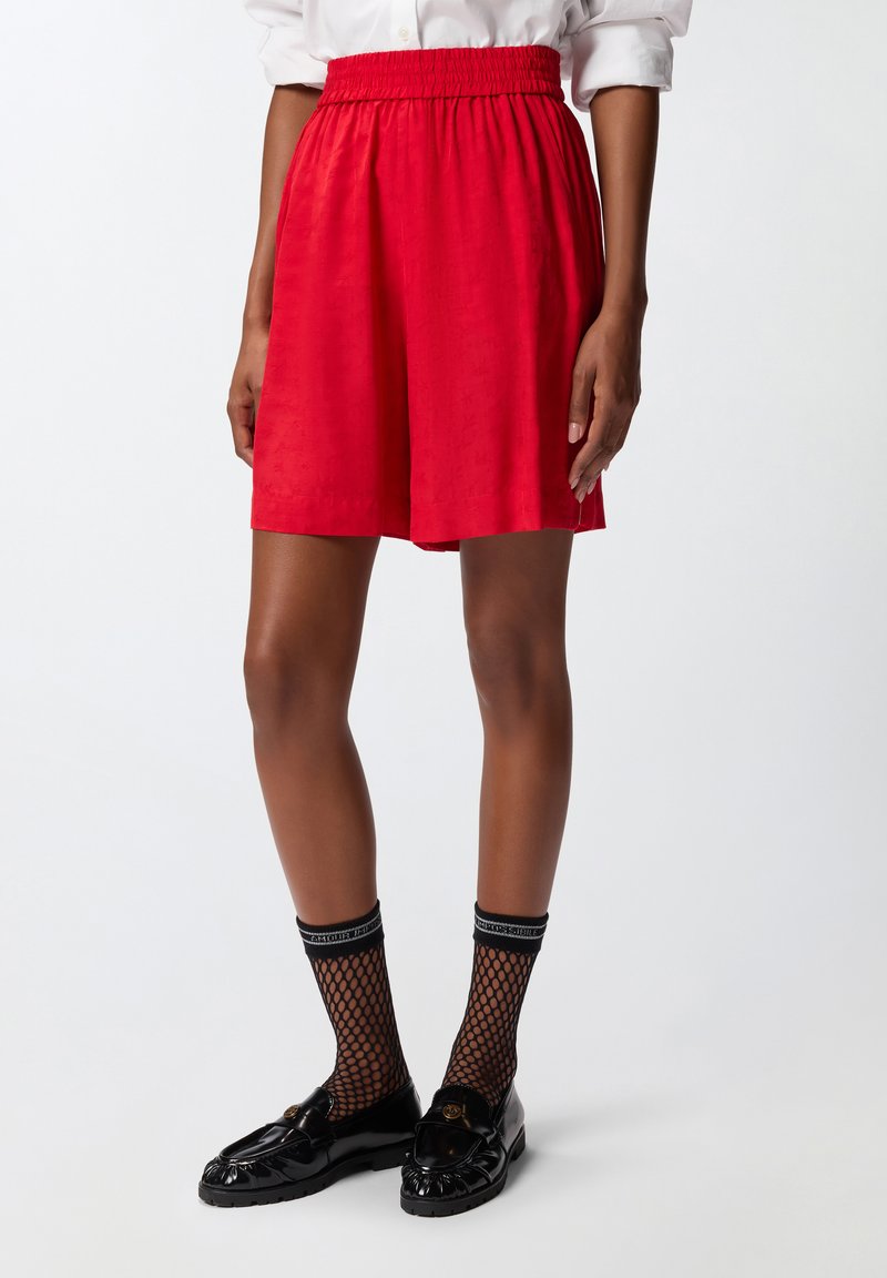 Shorts rossi con vita elastica, caratterizzati da una texture liscia e un motivo sottile. Abbinati a calze di rete nere e mocassini neri con un logo.