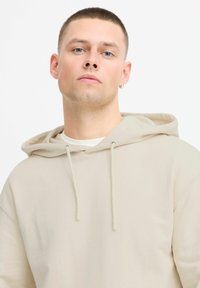 Hoodie color crema realizzato in morbido tessuto, con cappuccio regolabile, vestibilità comoda e design minimalista senza motivi visibili.