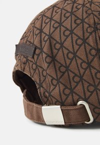 Brown fabric cap featuring a geometric black print, a leather label, and a metal adjuster with the brand name "Calvin Klein."