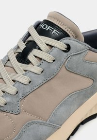 Baskets beige et gris en daim, avec des accents texturés, des lacets gris clair et un talon en cuir noir. Présente des détails de coutures et un logo.