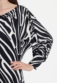Blouse noir et blanc avec un motif audacieux de rayures zèbre, coupe ample et manches froncées. Le tissu semble lisse avec une texture légère.