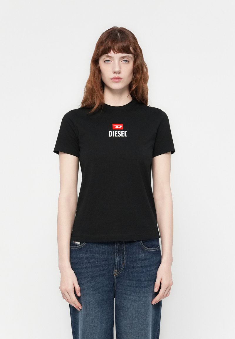 Donna con pelle chiara e capelli castani indossa una t-shirt nera Diesel e jeans blu, in piedi dritta contro uno sfondo bianco semplice.