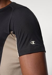 Μαύρο και καφέ αθλητικό μπλουζάκι με υφή υφάσματος, κοντά μανίκια και λογότυπο στο μανίκι. Ο σχεδιασμός είναι εφαρμοστ ός.