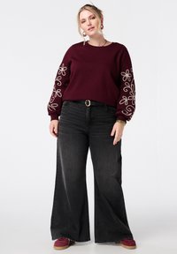 Bordeaux sweatshirt met bloemenborduursel op de mouwen, gecombineerd met zwarte wijde spijkerbroek en bordeauxkleurige schoenen. Eenvoudige ronde riem als accent.