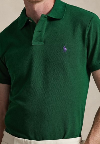 Πράσινο πόλο μπλουζάκι από υφασμάτινη ύφανση, με μοβ λογότυπο στο αριστερό στήθος, κλασικό γιακά και πατιλέτα με δύο κουμπιά.