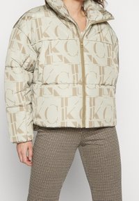 Puffjacka i beige med logomönster, med hög krage och dragkedja. Bärs tillsammans med rutiga byxor för kontrast.