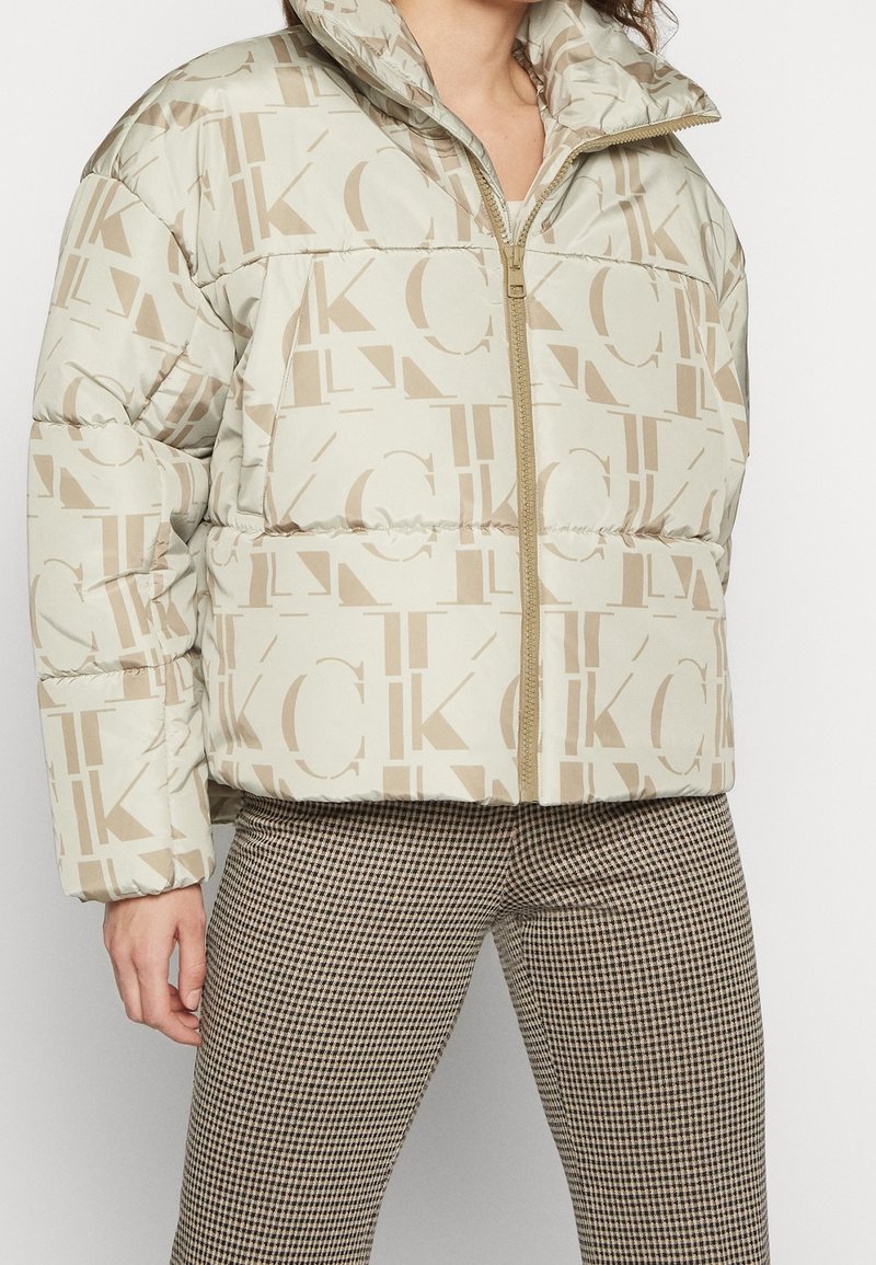 Puffjacka i beige med logomönster, med hög krage och dragkedja. Bärs tillsammans med rutiga byxor för kontrast.