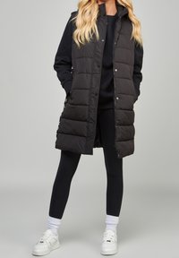 Black puffer vest with a hood, snap button closure, and quilted pattern, worn over a black outfit, paired with white sneakers and ribbed ankle socks.