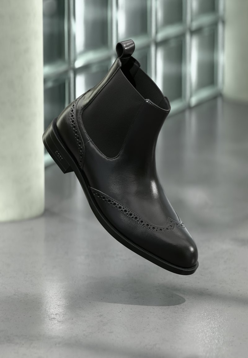 Cizmă Chelsea din piele neagră cu detalii brogue, panouri laterale elastice, tab pentru tragere și toc mic, prezentată în aer, pe fundalul unui podea de interior modern.