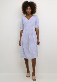 Striped blue and white dress with a V-neck, puff sleeves, and a flared hem. Smooth fabric falls to mid-calf. Worn with neutral slip-on shoes.