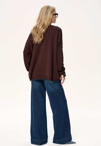 Brązowy, oversize'owy sweter w połączeniu z szerokimi dżinsami o kroju szerokich spodni. Sweter ma fakturowane wykończenie, a dżinsy są głęboko niebieskie z podwiniętymi nogawkami.