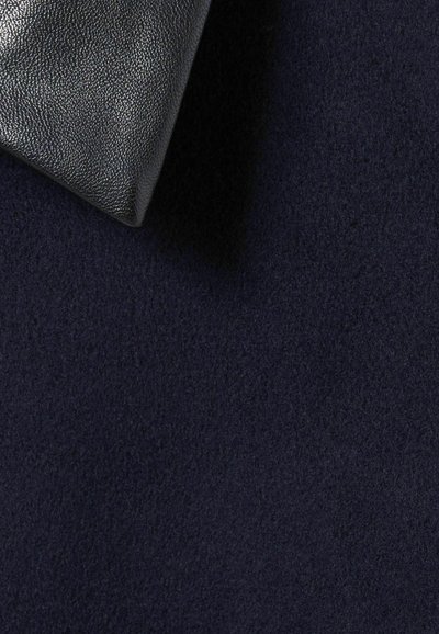 Funda de cuero negro texturizado con forma puntiaguda, reposando sobre un fondo de tela azul marino oscuro con una textura suave y lisa.