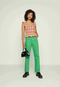 Mulher jovem em pé, vestindo uma blusa sem mangas com volantes multicoloridos, calças verdes, sapatos de plataforma pretos e segurando uma pequena bolsa de mão preta.