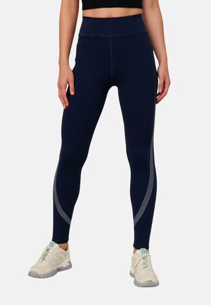 Žena nosi tmavomodré legíny s vysokým pásom s bielym zakriveným pruhom a béžové tenisky so zelenými akcentmi.